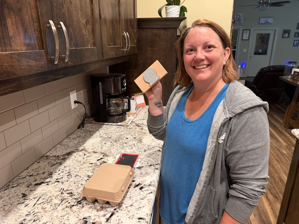 Smiling Aragon Acres co-owner proudly holding the farm's wooden logo stamp beside a branded egg carton.