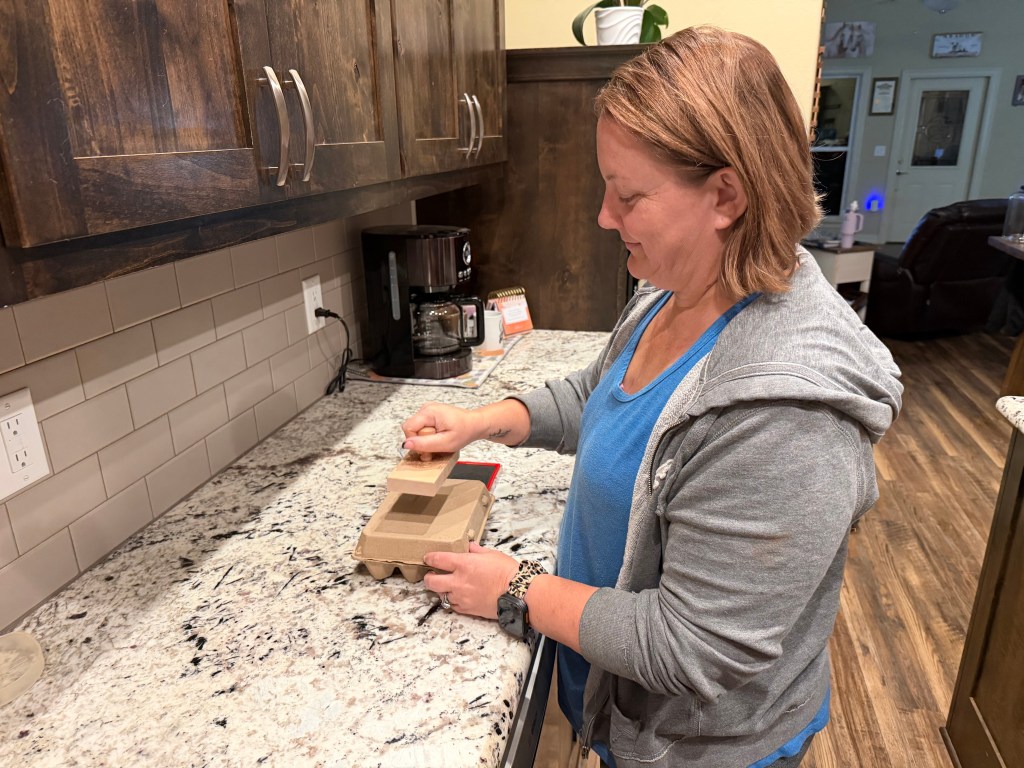 Stamping the Aragon Acres farm logo onto a cardboard egg carton using a wooden stamp on the kitchen counter.